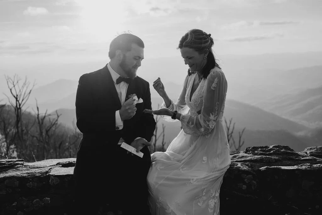 Black and white close-up of groom placing wedding ring on bride's finger during intimate elopement ceremony at Wayah Bald summit, North Carolina