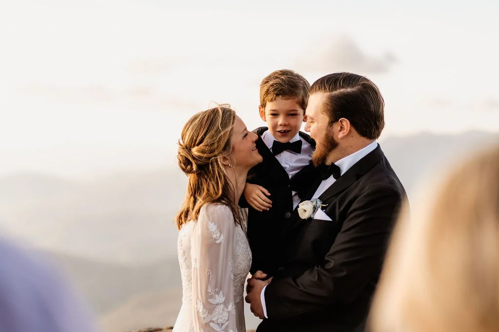 Bride, groom, and young son laughing joyfully together on the Wayah Bald summit at golden hour during their intimate family mountain elopement, Nantahala NC