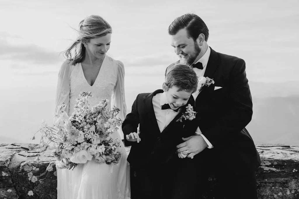 Black and white photo of bride, groom, and son in tuxedo laughing together during their intimate mountaintop wedding celebration at Wayah Bald, North Carolina