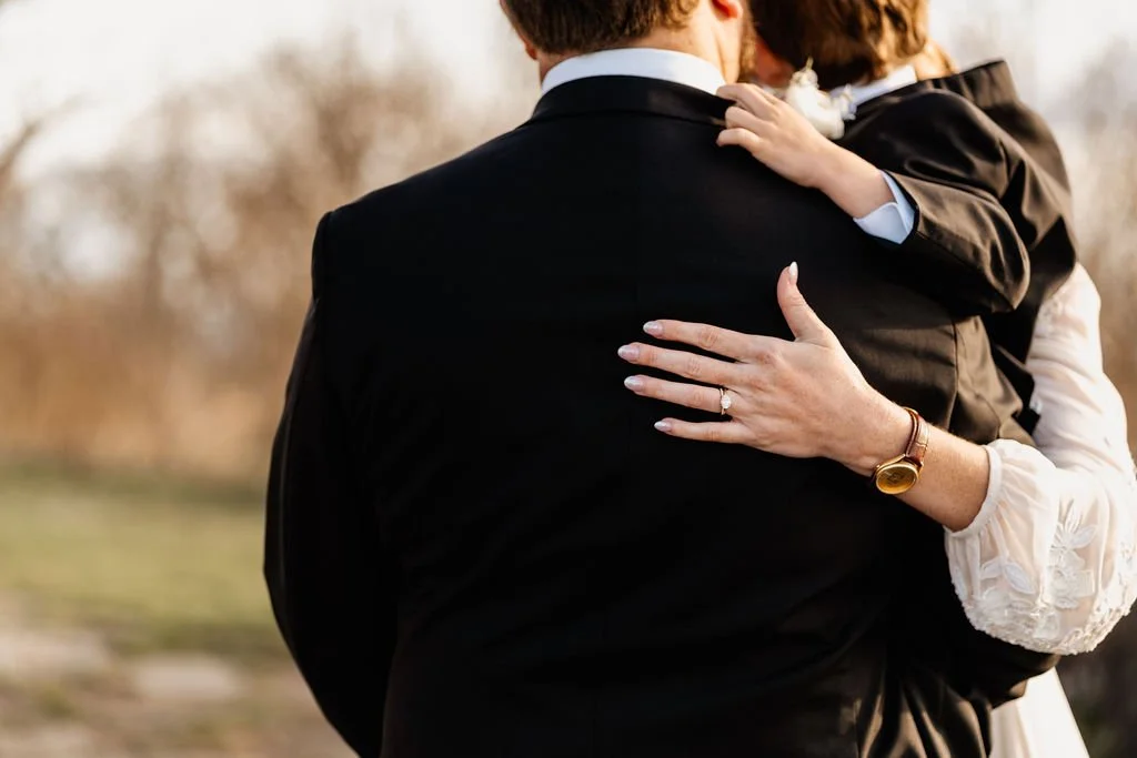 Close-up from behind of bride and groom embracing after their intimate wedding ceremony at Wayah Bald, bride's diamond ring and embroidered lace gown detail visible