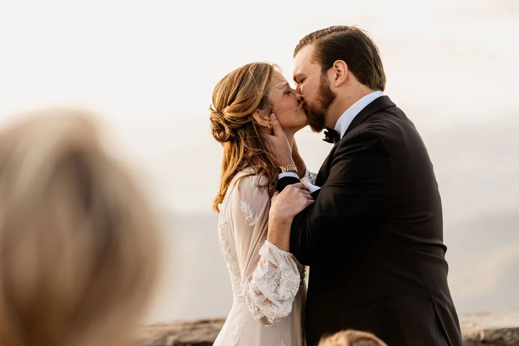 Bride and groom sharing their first kiss as a married couple at Wayah Bald summit with glowing golden hour Appalachian mountain ridges behind them, Nantahala NC