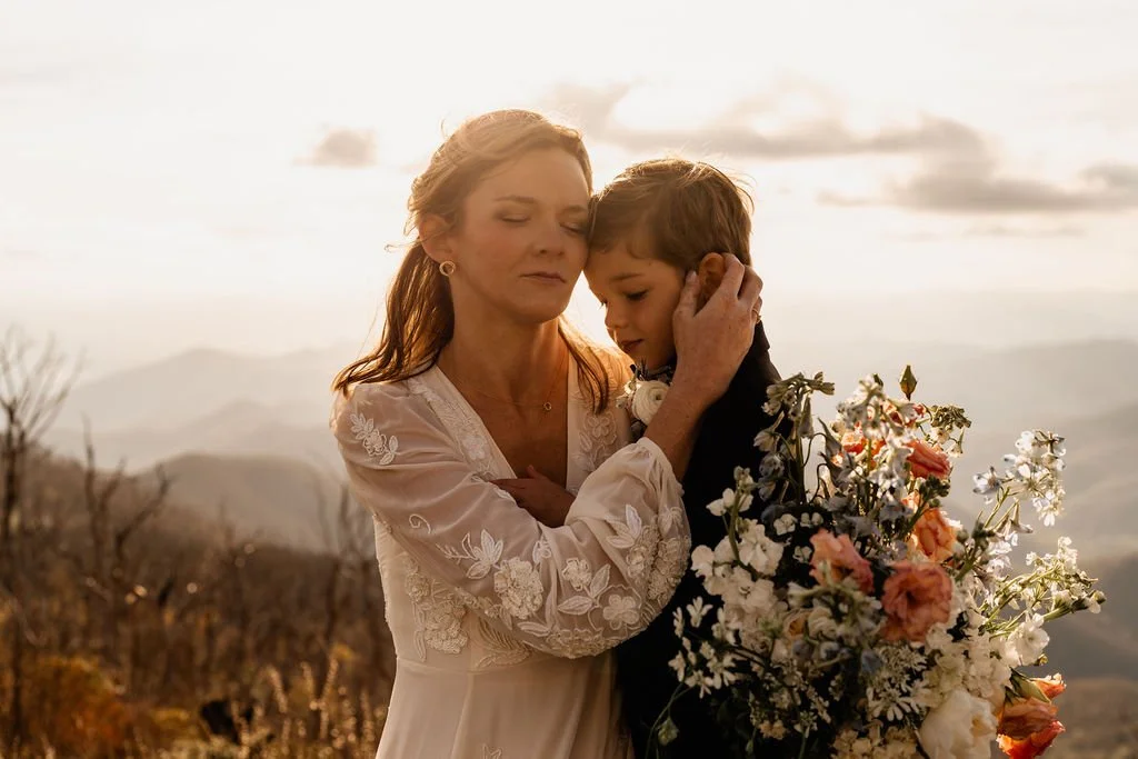 Bride in lace wedding gown tenderly embracing her young son in a warm golden-light moment during their Wayah Bald mountain elopement, Nantahala National Forest NC