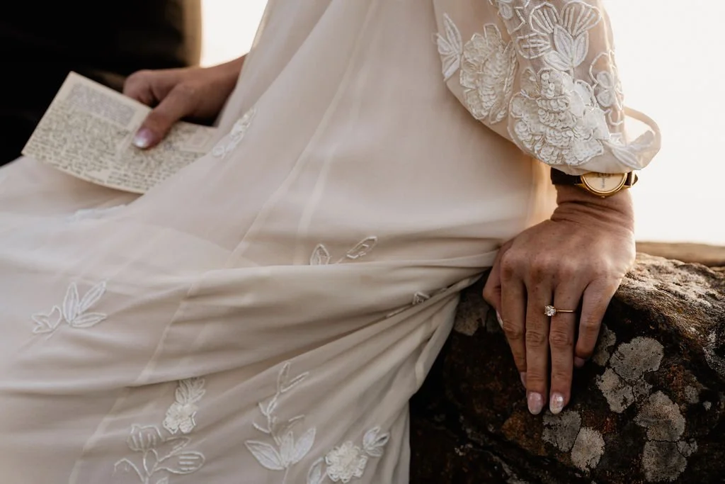Close-up of diamond engagement ring resting on bride's embroidered lace wedding gown sleeve against the stone surface at Wayah Bald elopement, Nantahala National Forest NC