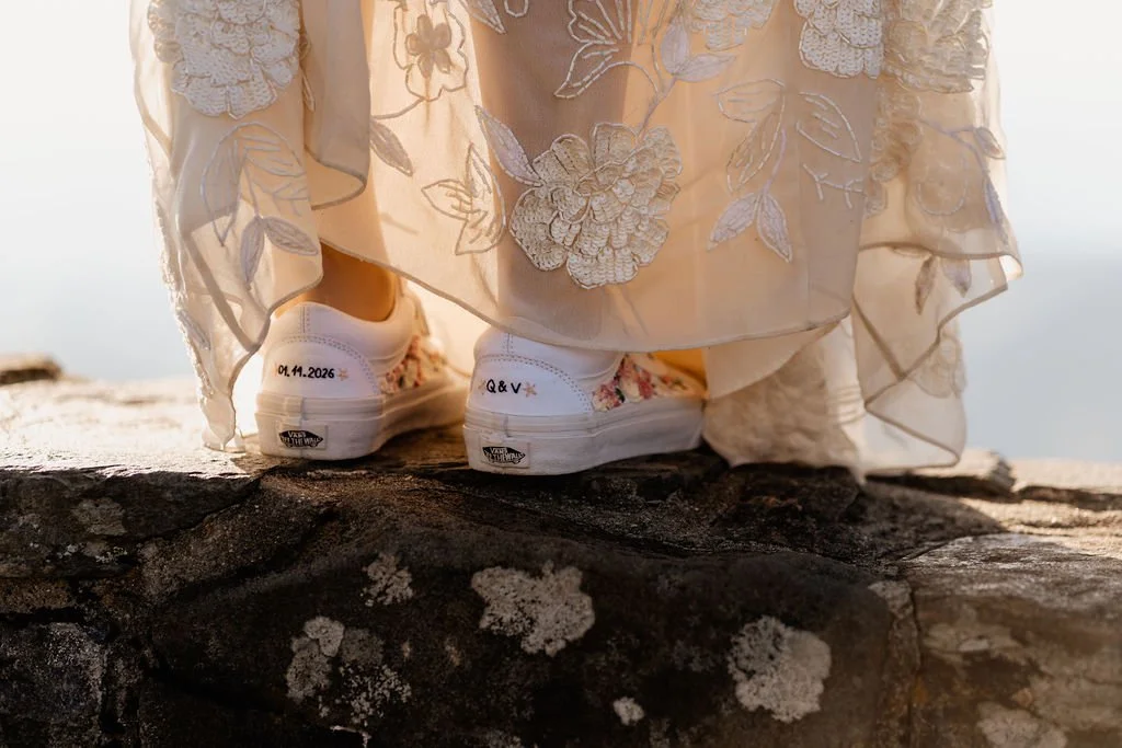 Close-up of bride's white sneakers with wedding date written in gold script on the sole, resting on stone surface at Wayah Bald during intimate mountain elopement, NC