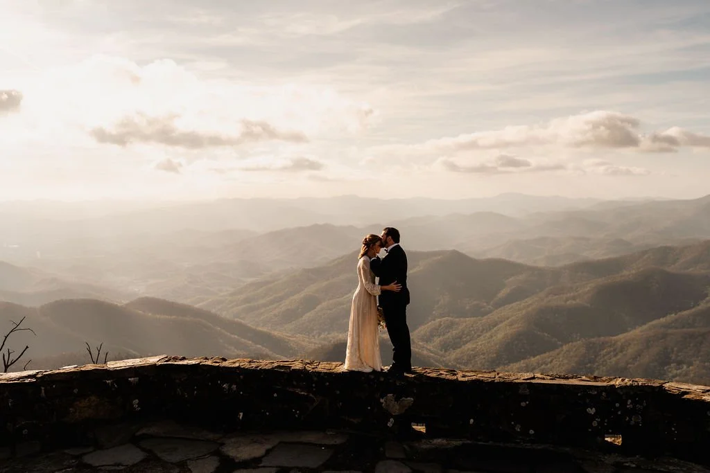 Silhouette of bride and groom sharing a kiss on the Wayah Bald stone overlook ridge wall at golden sunset with dramatic Appalachian mountain panorama behind them