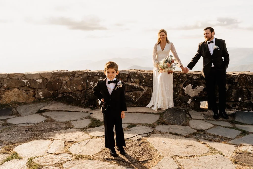 Family portrait of bride, groom, and young son on the stone terrace at Wayah Bald summit at golden hour during their intimate mountain wedding, Nantahala NC