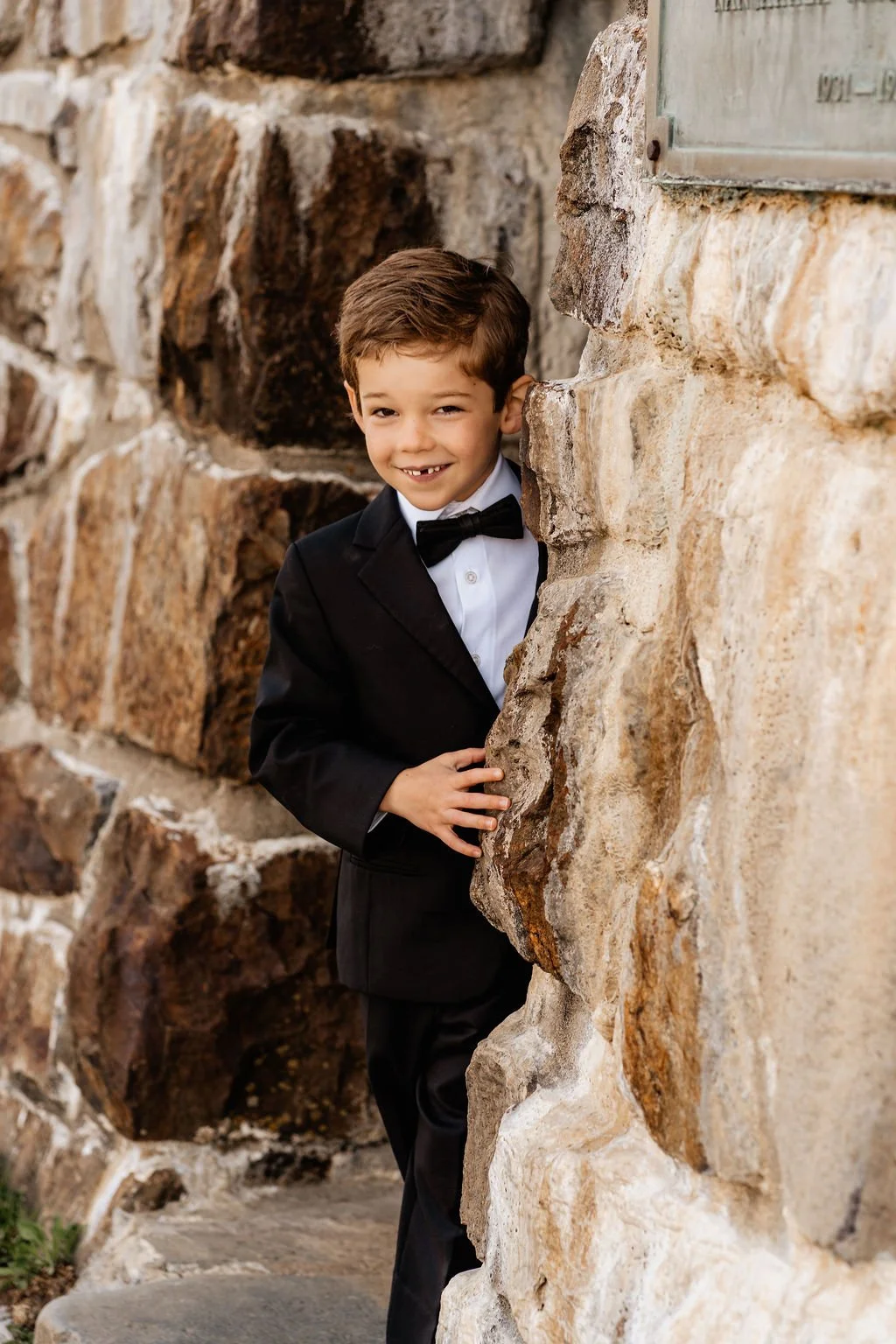 Young boy in tuxedo smiling and peeking playfully around the corner of the Wayah Bald stone tower wall during his parents' intimate Appalachian mountain elopement