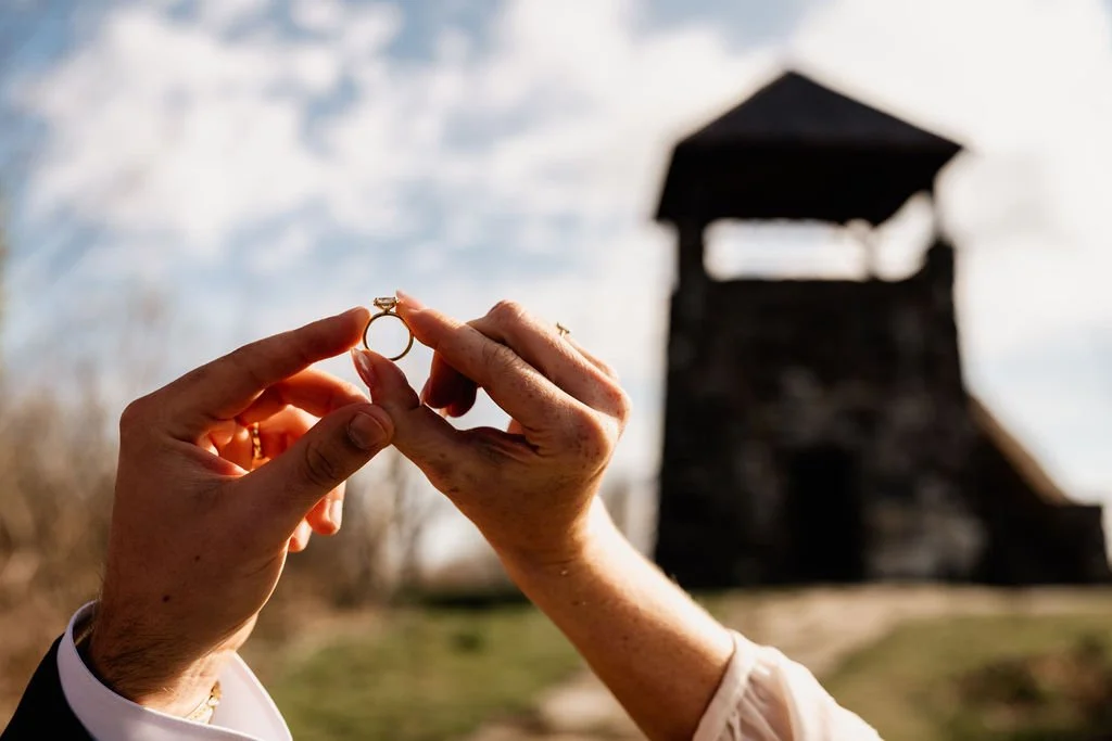Close-up of bride holding up her diamond wedding ring against the sky with the Wayah Bald historic stone fire tower blurred in the background, Nantahala National Forest NC