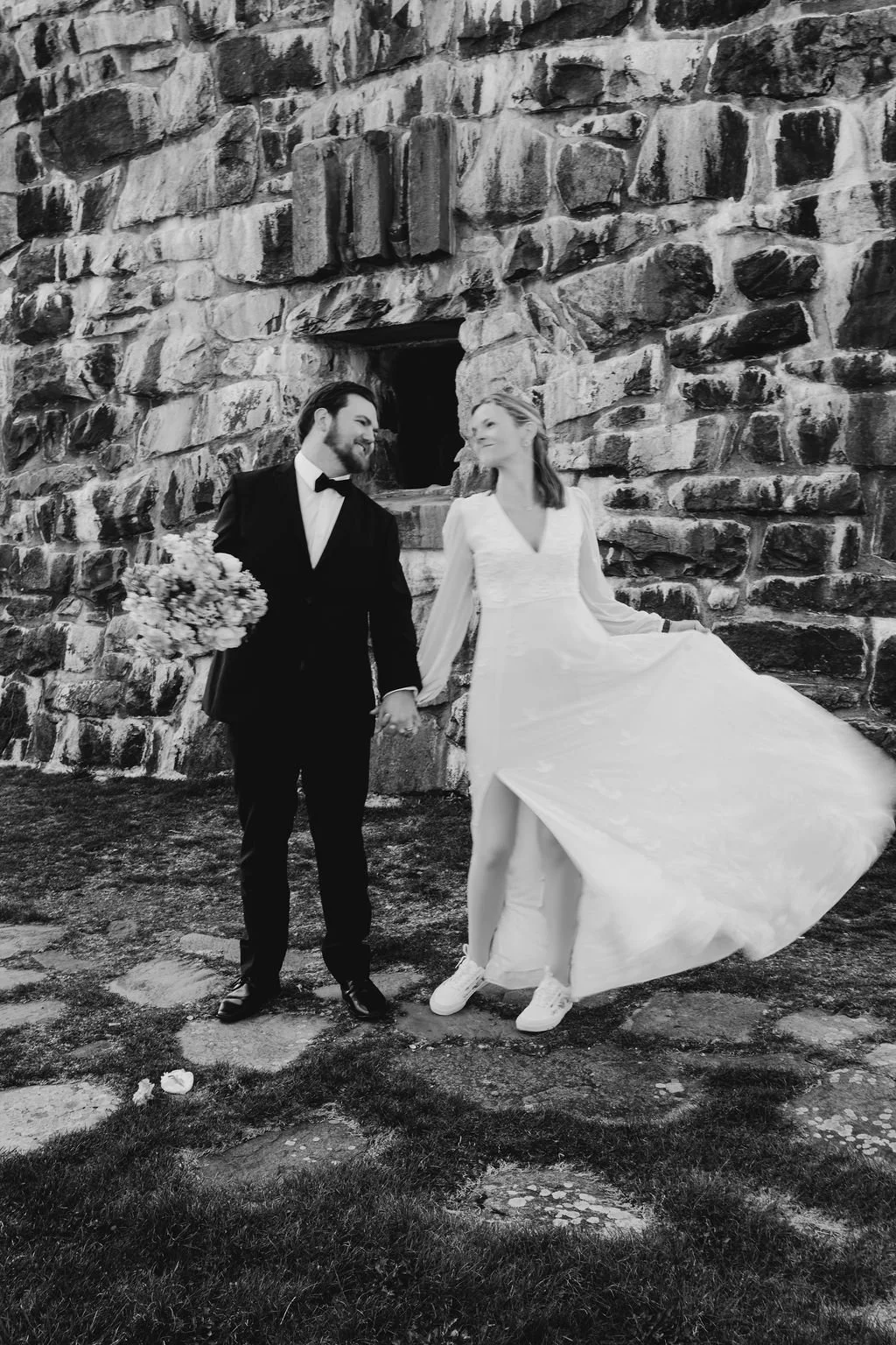 Black and white portrait of bride and groom standing hand in hand in front of the Wayah Bald historic stone fire tower, bride's wedding gown flowing in the wind, Nantahala NC