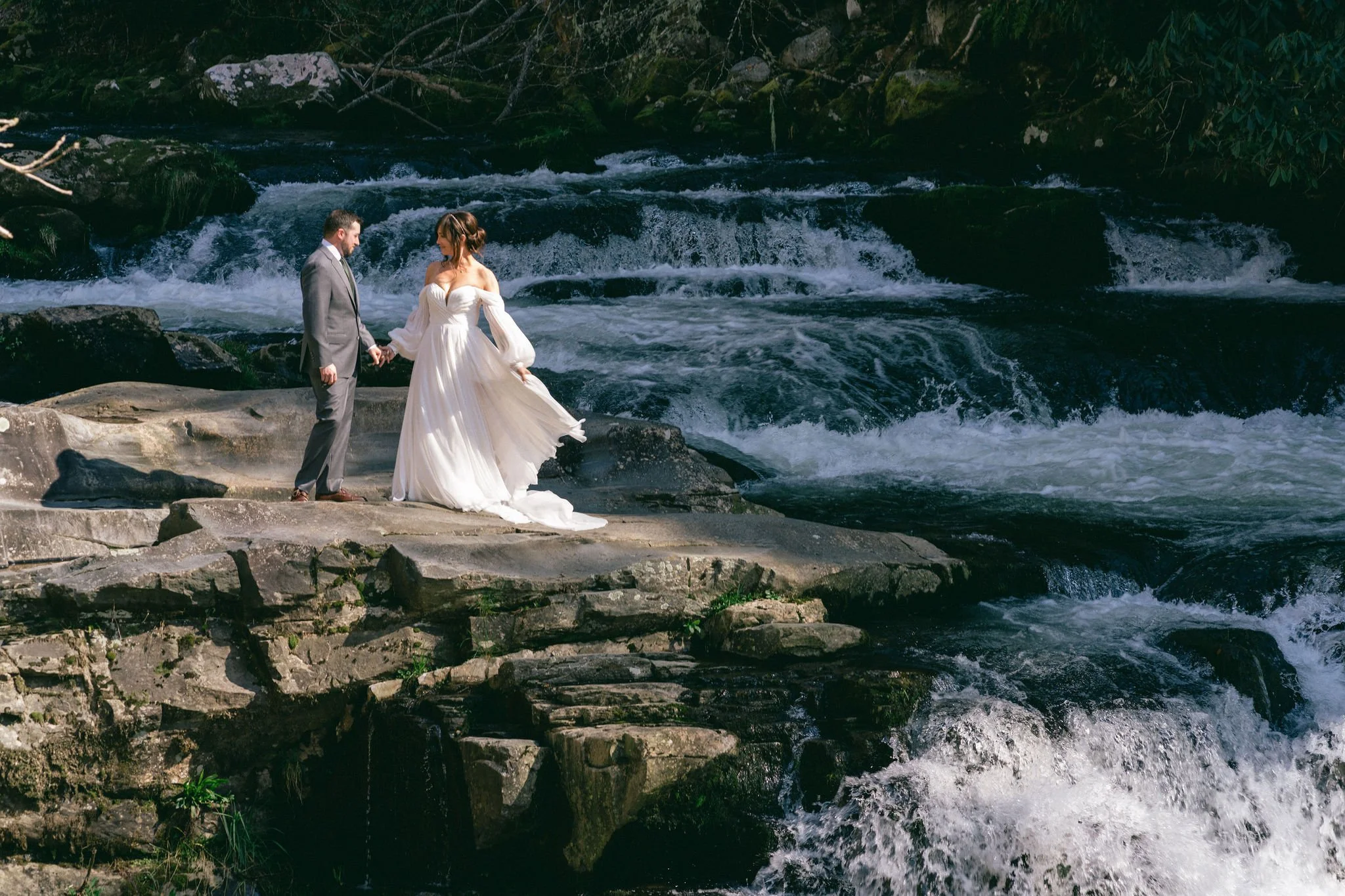Bride in flowing off-shoulder wedding gown holding hands with groom on rocky riverbank beside rushing Appalachian waterfall, Blue Ridge Georgia elopement photographer.jpg