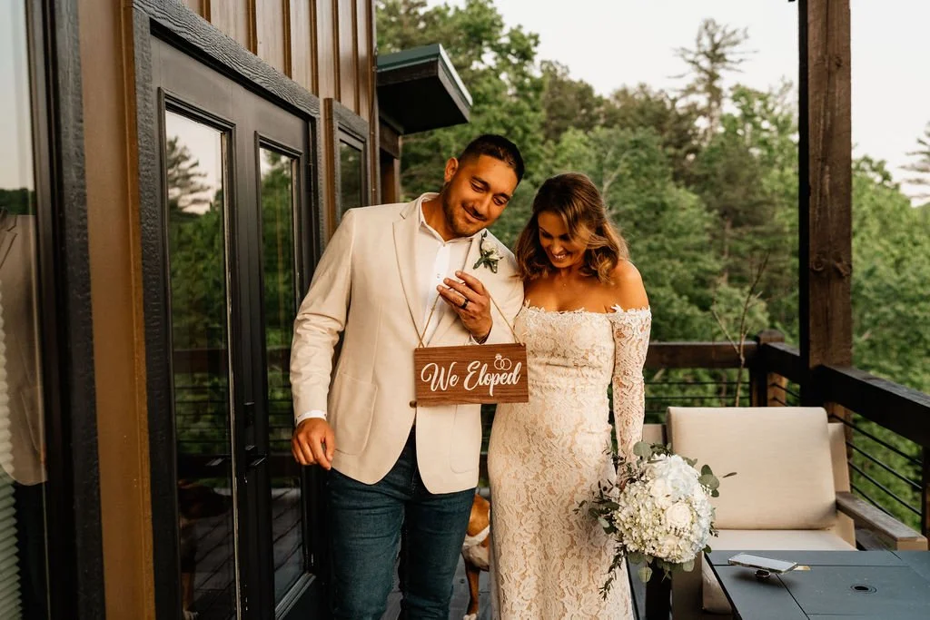 Smiling couple holding a wooden We Eloped sign on a cabin porch with Appalachian forest behind them, Blue Ridge Georgia elopement photographer.jpg