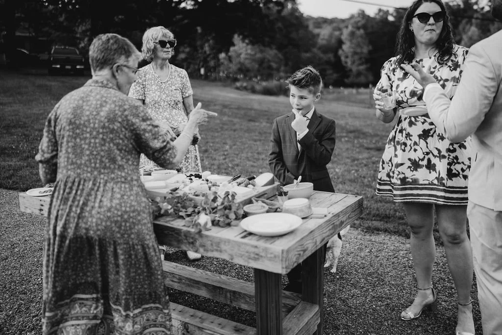 Black and white candid photograph of wedding guests and a young boy at an outdoor grazing board during a Blue Ridge, Georgia barn wedding reception