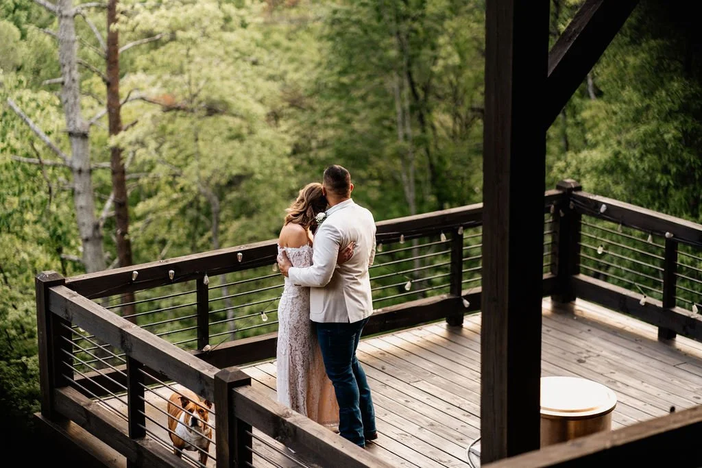Bride and groom embracing on a cabin deck overlooking lush Appalachian forest with their dog nearby, intimate Blue Ridge Georgia cabin elopement, Appalachian Wedding Photography