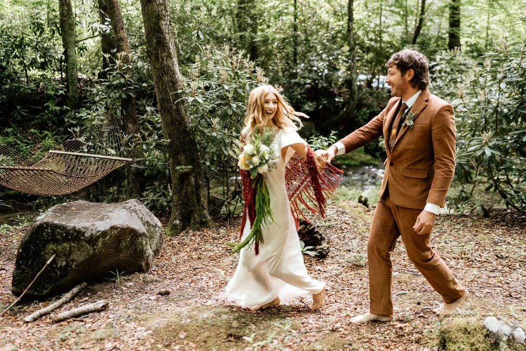 Hope and Chase walking joyfully through the Appalachian forest at Huffman Creek, bride carrying a red fringe bouquet, groom in a rust suit
