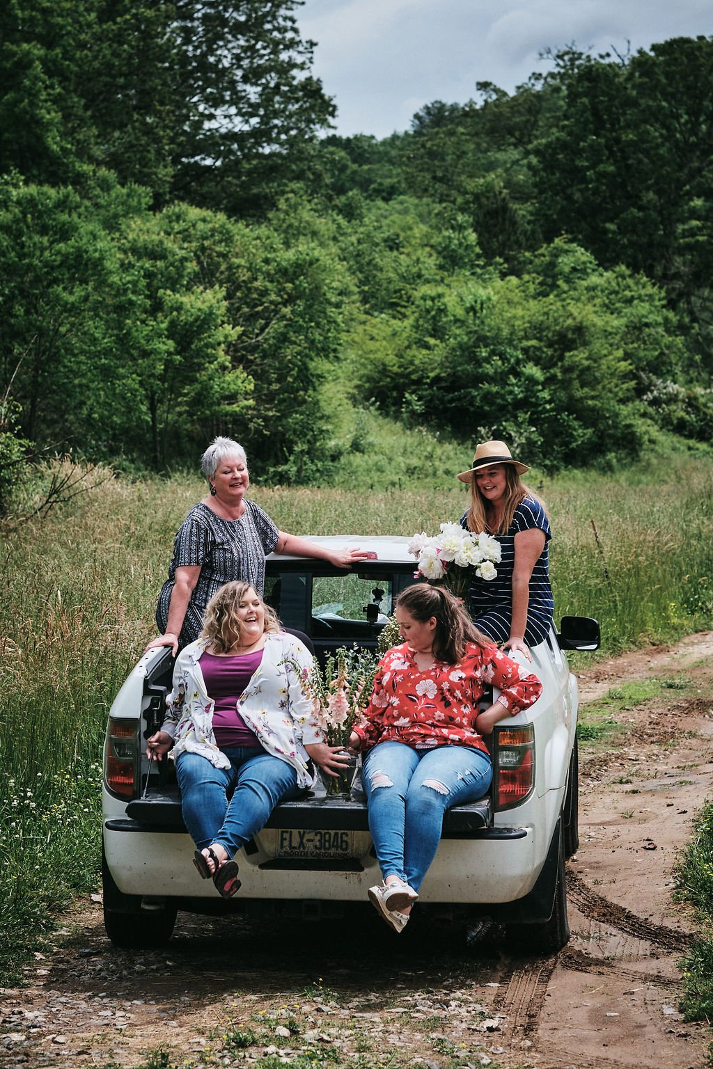Four women gathered around the open tailgate of a white pickup truck on a forest dirt road in Blue Ridge Georgia, holding wildflower bouquets