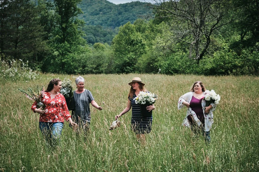 Four women walking through a tall-grass meadow carrying armfuls of wildflowers with Blue Ridge Georgia mountains in the background