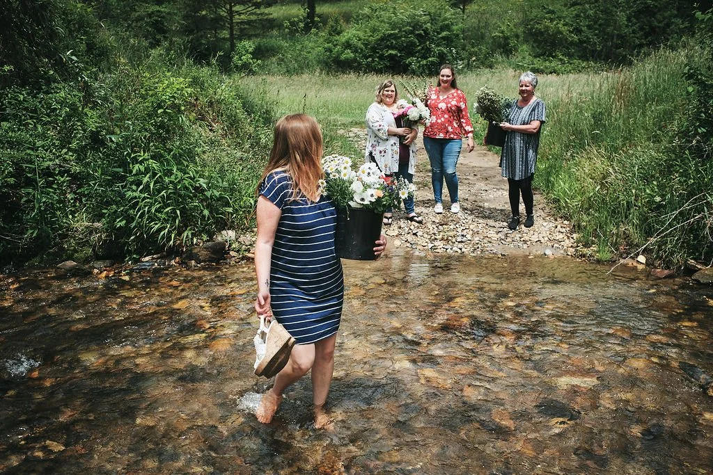 Woman wading barefoot through a shallow mountain creek holding her sandals while three women watch from the bank in Blue Ridge Georgia