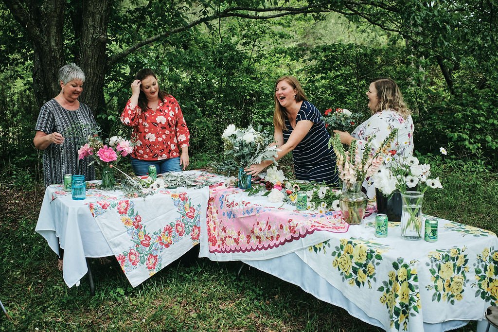 Four women laughing while arranging flowers around an outdoor table covered in vintage floral tablecloths in a Blue Ridge Georgia forest clearing
