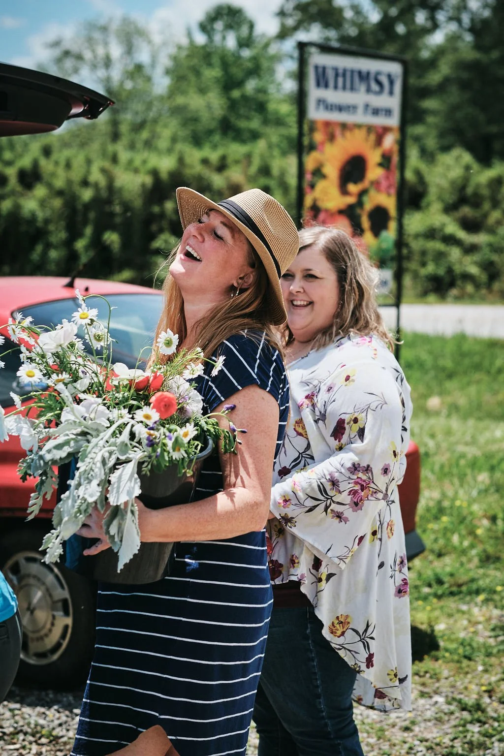 Two women laughing outside Whimsy Flower Farm holding a pot of wildflowers with the farm's sunflower sign visible in Blue Ridge Georgia