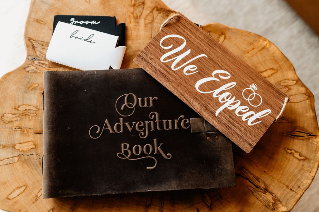 Flat-lay of elopement details on a wood slice including an "Our Adventure Book" leather journal, "We Eloped" wooden sign, and bride and groom vow cards from Erin and William's Blue Ridge Georgia