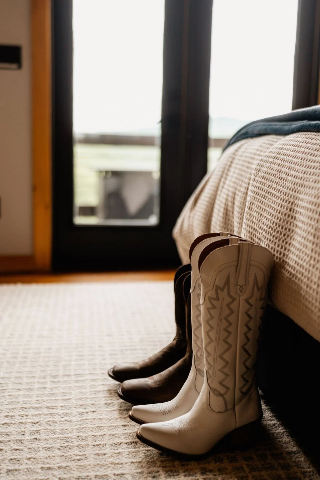 Erin's white embroidered cowboy boots and William's brown cowboy boots standing side by side on the cabin floor with a mountain view through the glass door behind them