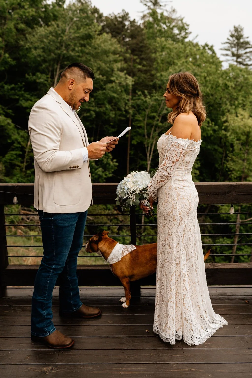 William reading his vows to Erin on the cabin deck with their dog standing between them in a lace bandana and the Appalachian forest behind them during their Blue Ridge Georgia elopement ceremony