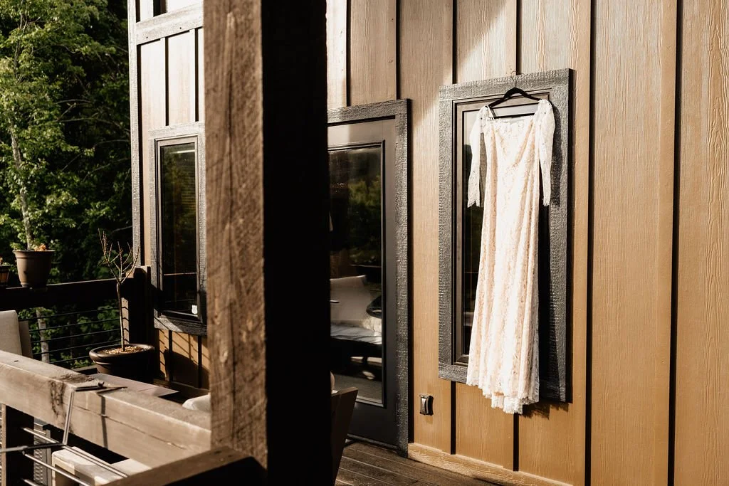 Erin's lace wedding gown hanging on a hanger on the dark wood exterior of a Blue Ridge Georgia cabin, morning light illuminating the lace detail