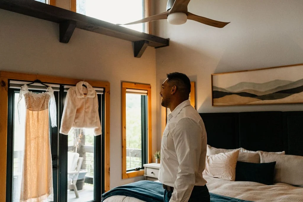 William standing in a cabin bedroom looking toward Erin's lace wedding gown and white fur wrap hanging by the window during their Blue Ridge Georgia elopement