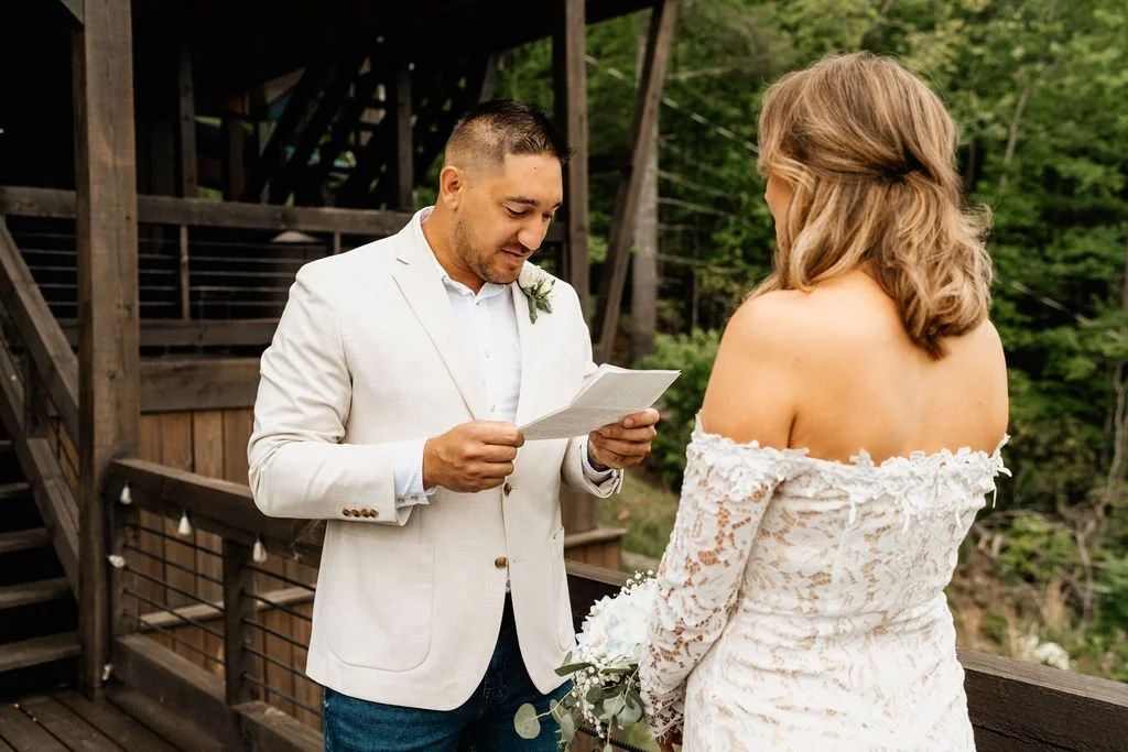 William reading his personal vows from a piece of paper on the cabin deck in his cream blazer, smiling, during their Blue Ridge Georgia elopement ceremony