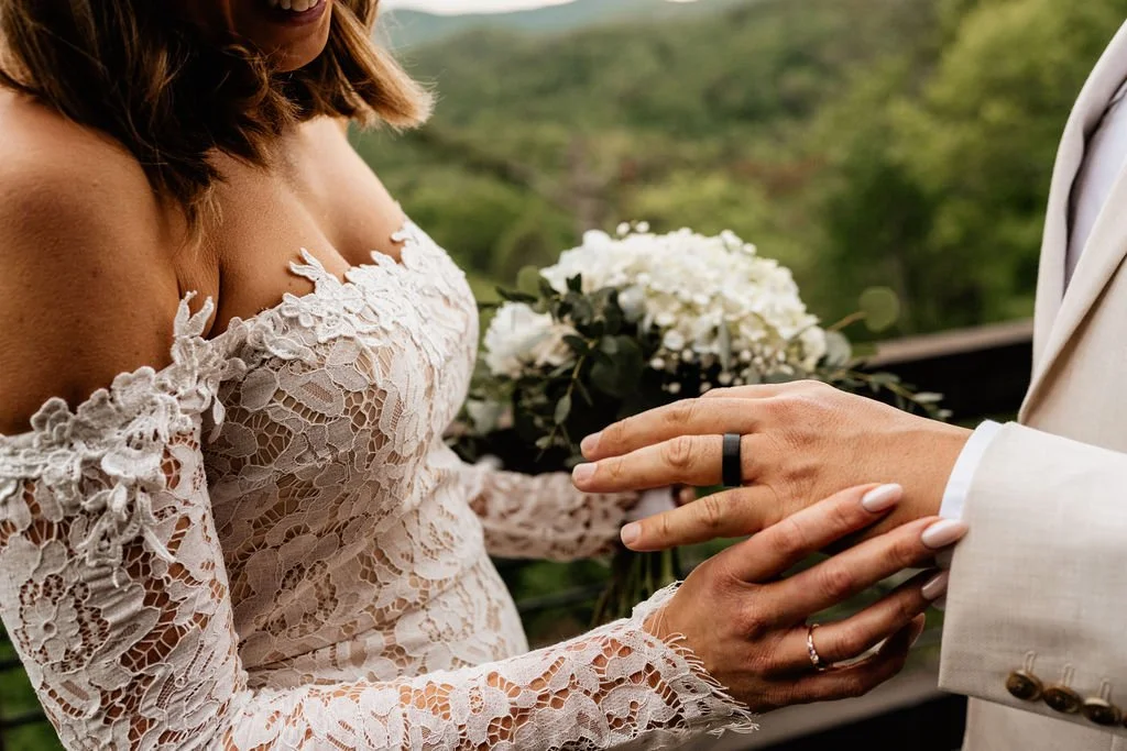 Close-up of William placing a black wedding band on Erin's finger during their ring exchange on the cabin deck, with blue hydrangea bouquet and Appalachian forest softly blurred behind them