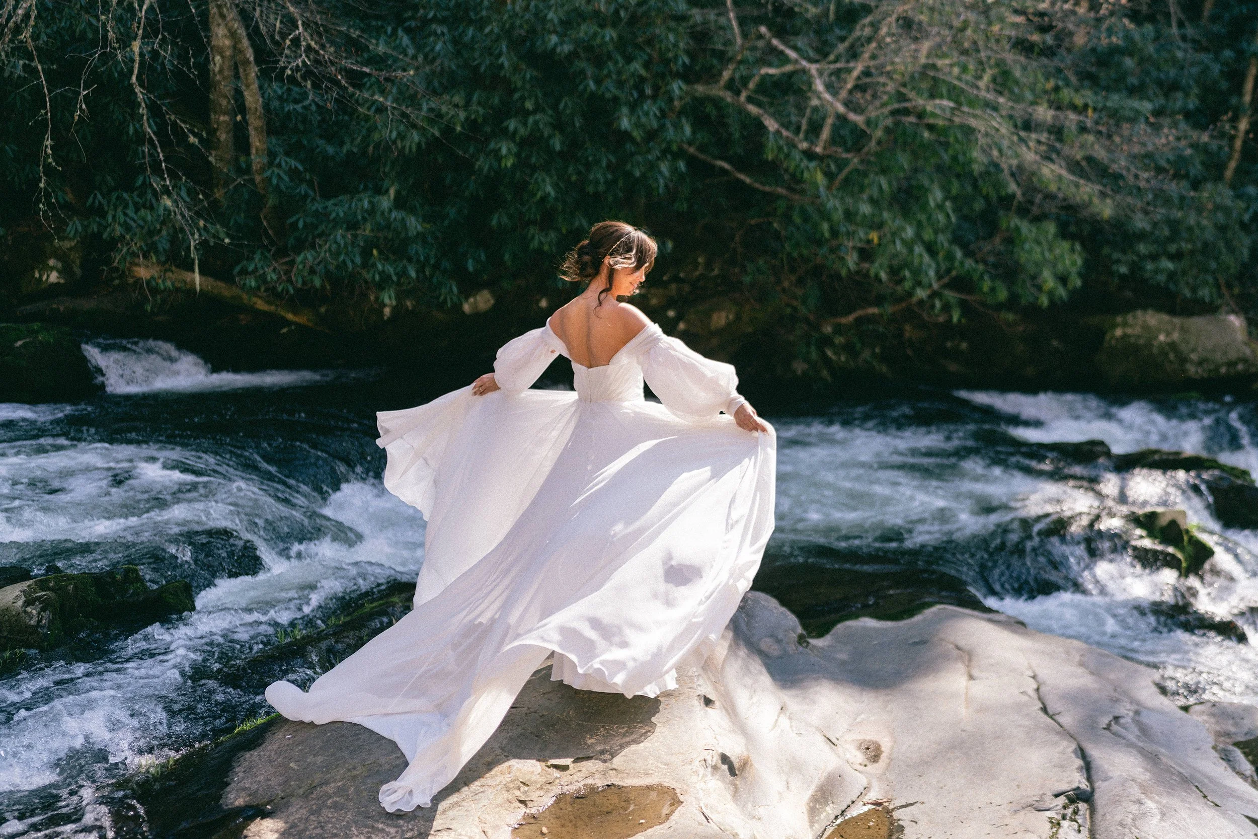 Bride in a flowing off-shoulder gown standing on river rocks with rushing water in the Appalachian mountains