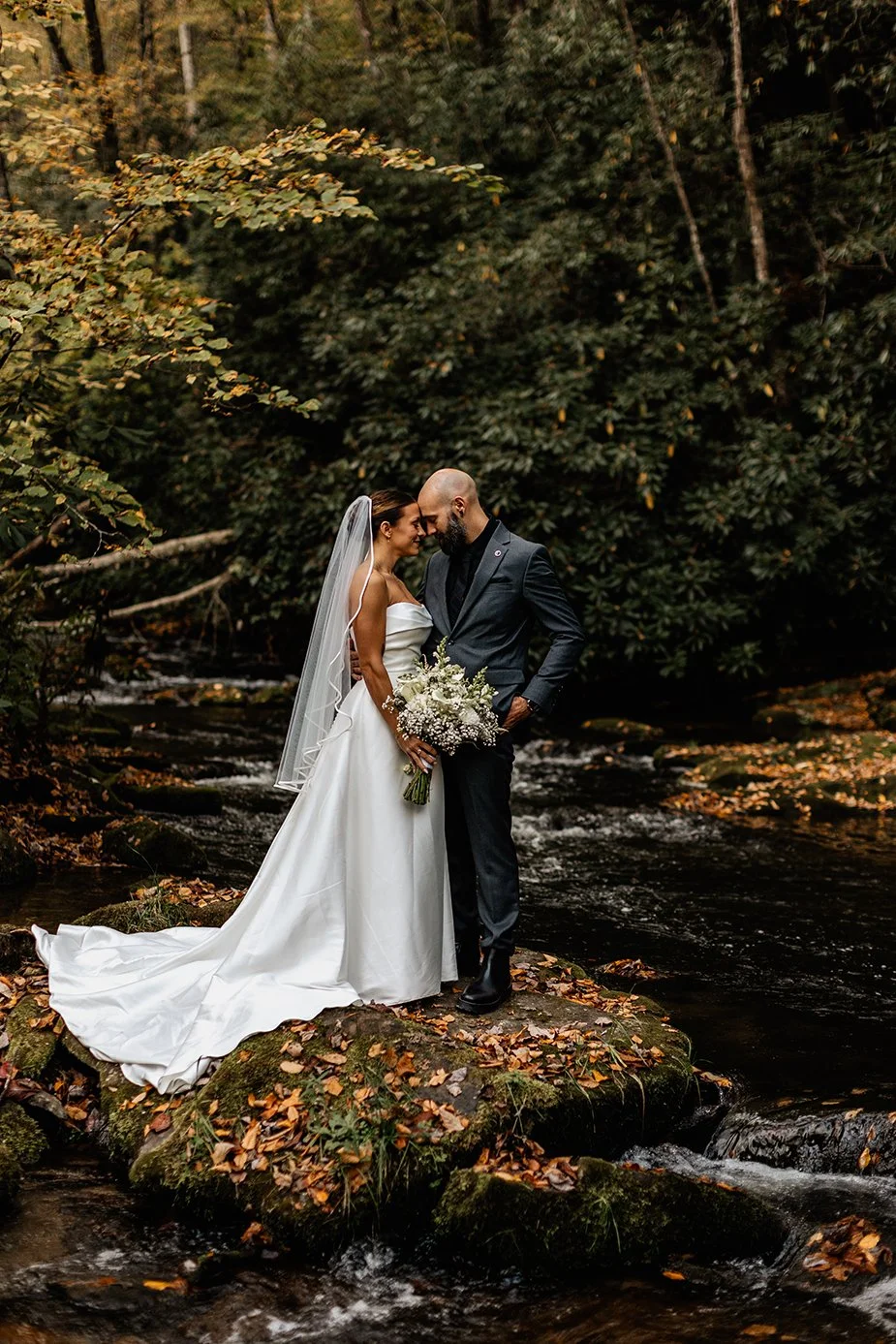 Couple touching foreheads tenderly on mossy rocks beside an autumn creek during their Appalachian elopement