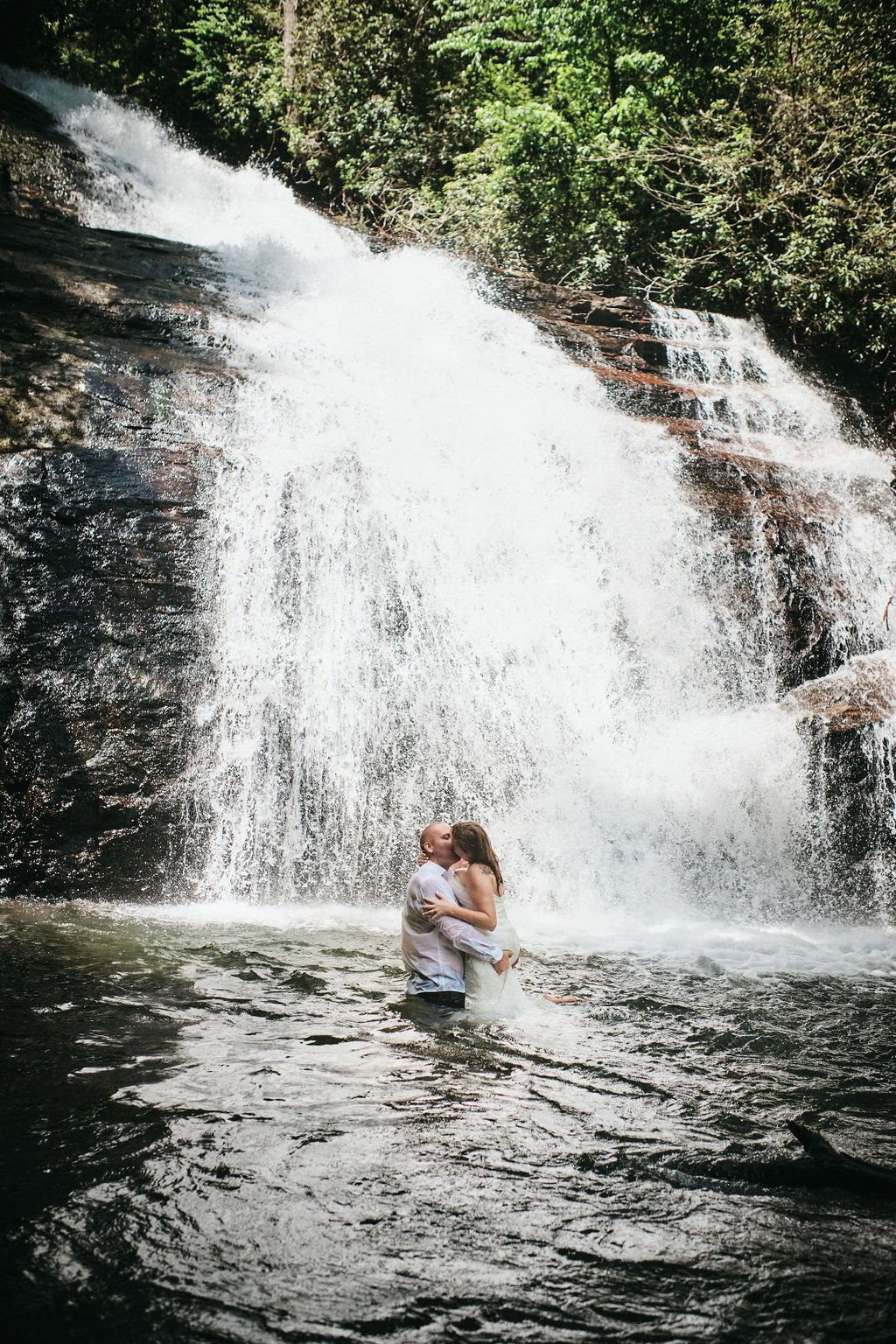 Vickie and Cody kissing waist-deep in the pool at the base of a cascading waterfall, both in soaked wedding attire, during their Blue Ridge Georgia elopement