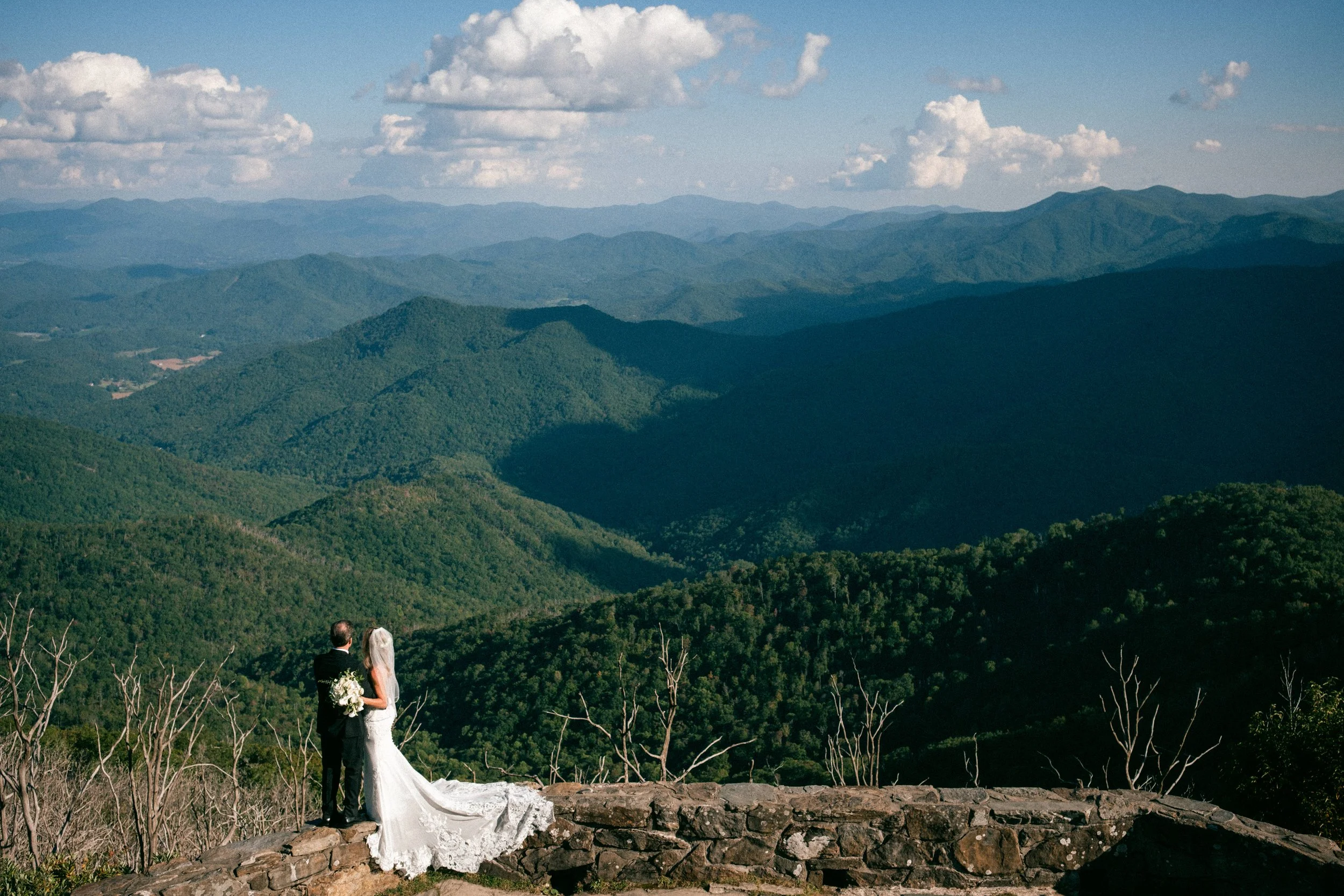 A bride and groom in wedding attire standing on a stone overlook with a mountain range and cloudy sky in the background.