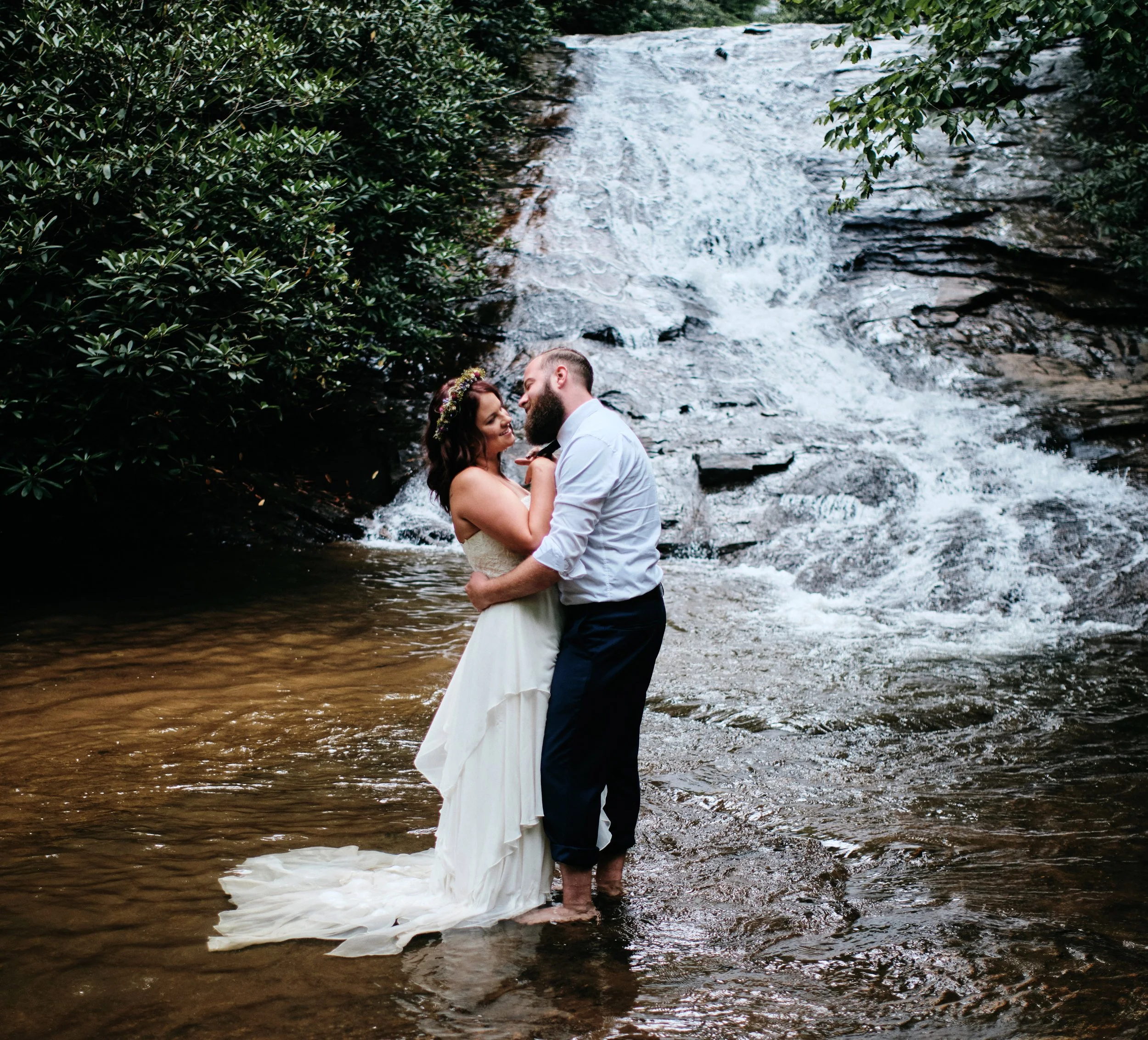 Bride and groom embracing barefoot in a creek in front of a waterfall during their wedding session