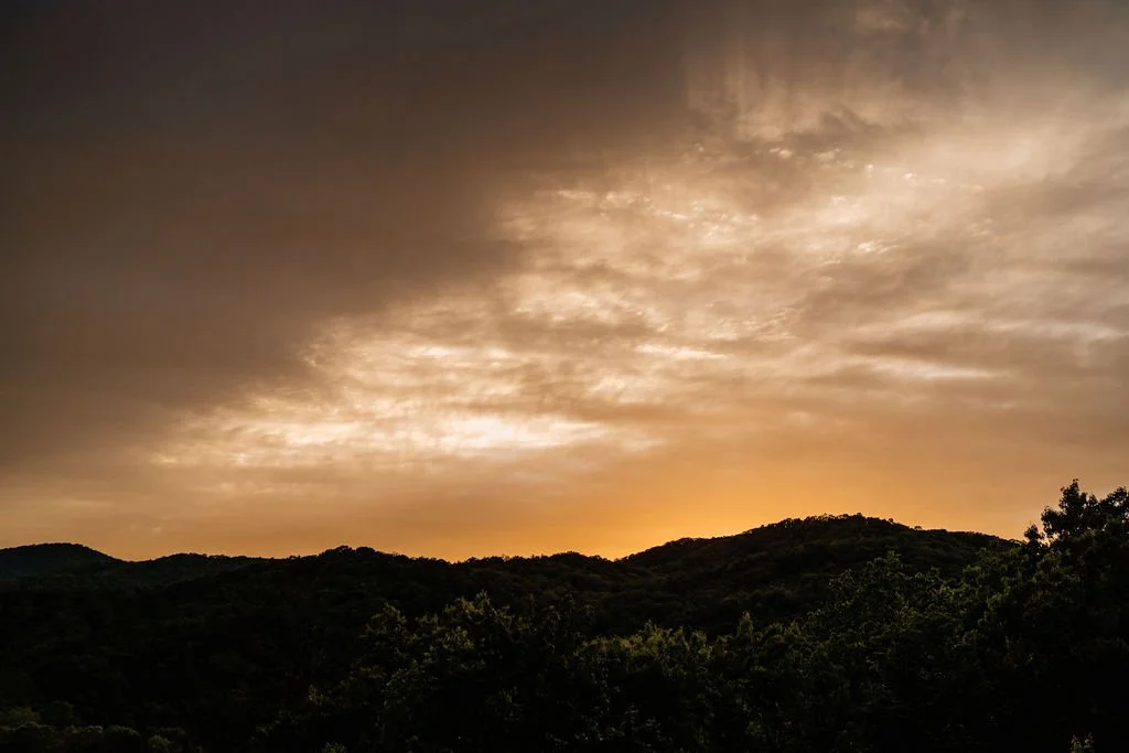 Dramatic Blue Ridge Georgia mountain sunset with deep orange glow on the horizon, layered mountain silhouettes, and storm clouds lit gold and amber, viewed from the cabin deck during Erin and William's elopement