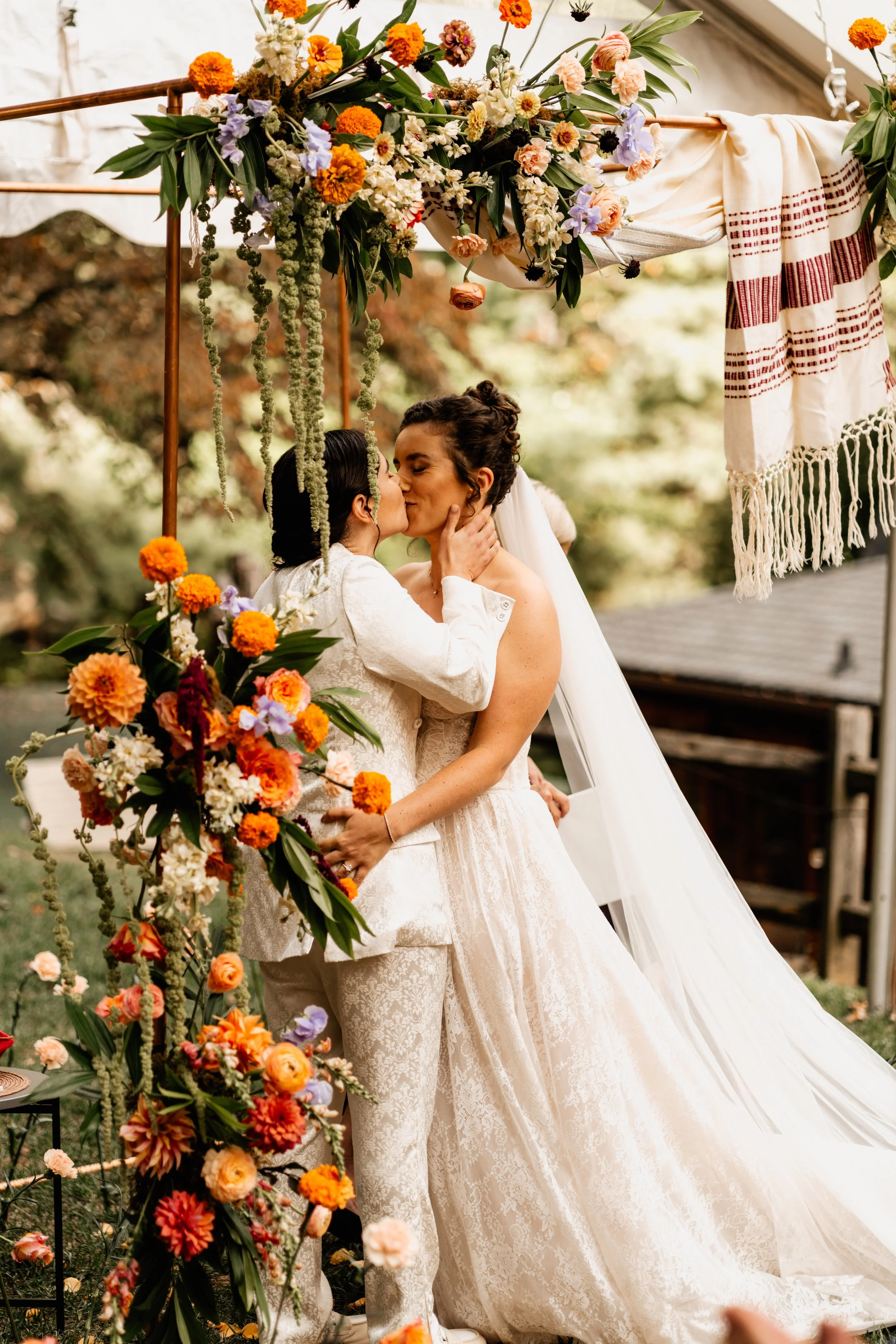 Two brides kissing under a floral wedding arch filled with orange flowers