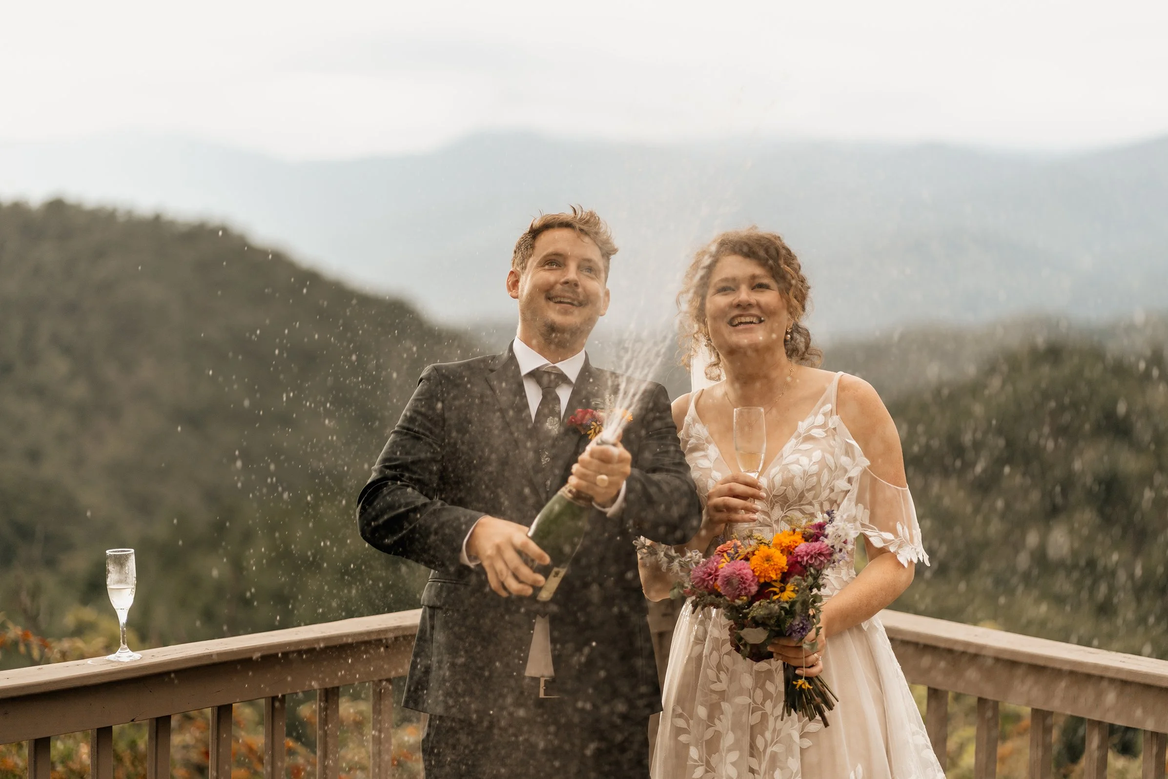 Greyson and Miranda laughing as champagne sprays on a mountain deck with misty Appalachian ridgelines in the background during their Blue Ridge wedding