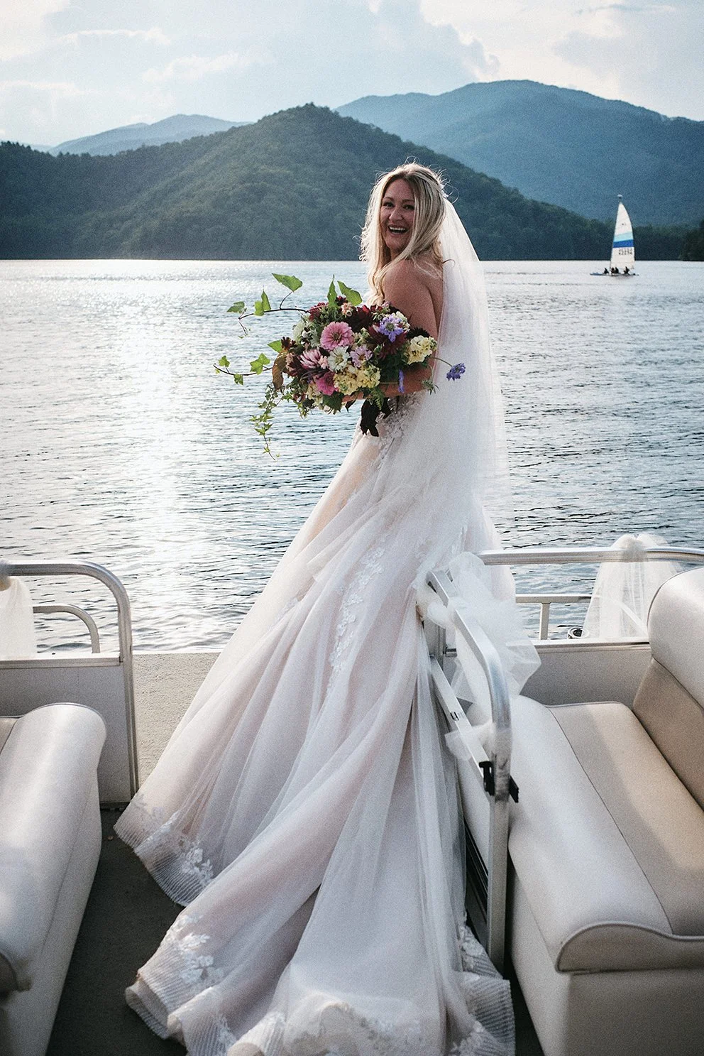 Bride in a flowing white gown seated on a pontoon boat on a tranquil mountain lake during an intimate Appalachian wedding
