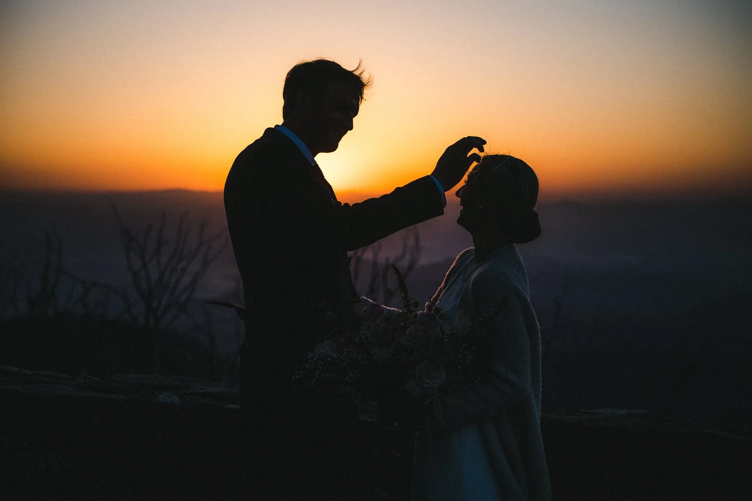 A silhouette of a man and woman during sunset, with the man touching the woman's forehead, creating an intimate moment.