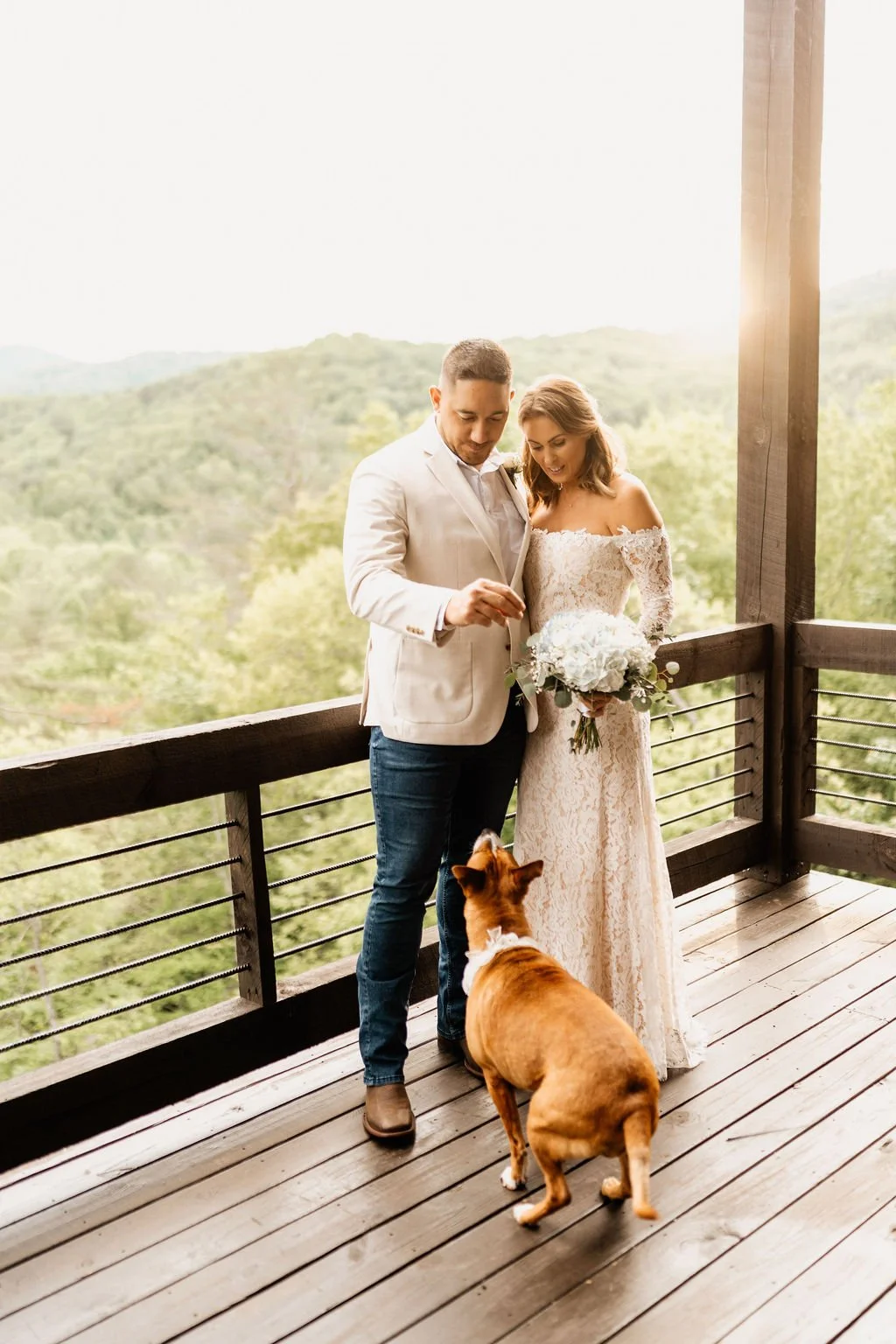 Erin and William standing on the cabin deck at golden hour with mountain ridgelines behind them and their dog in a lace bandana looking up at them, Blue Ridge Georgia elopement