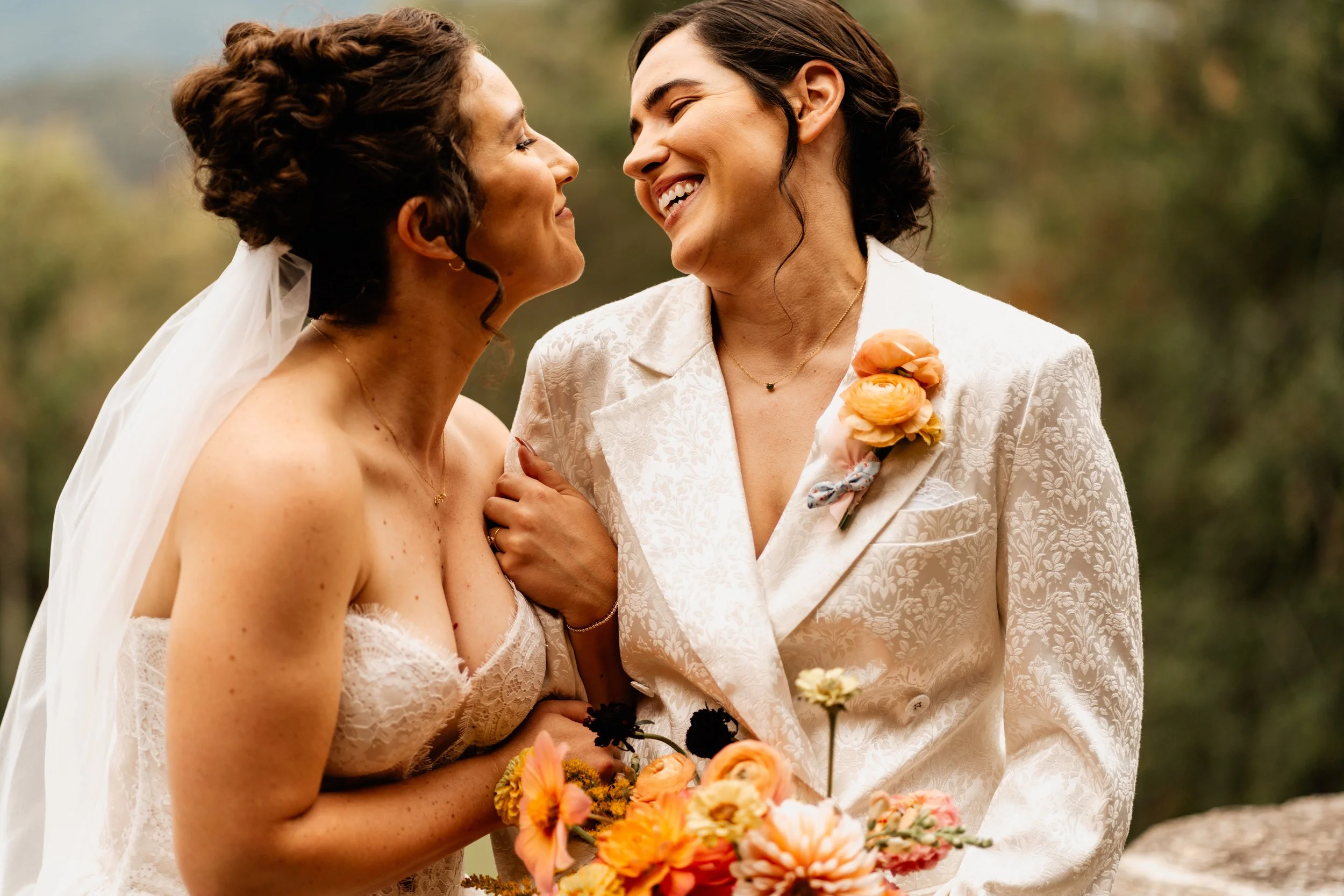 Maddie and Brenna laughing nose to nose on their wedding day, one in a white suit and one in a veil with an orange bouquet