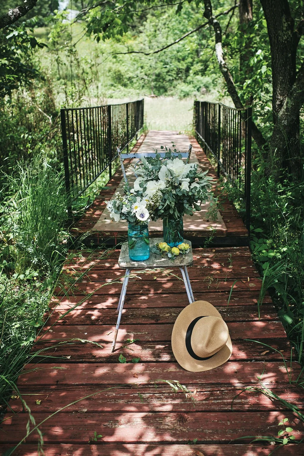 Two mason jar vases of white wildflowers on a vintage stool with a straw hat resting on a rustic red wooden bridge in Blue Ridge Georgia
