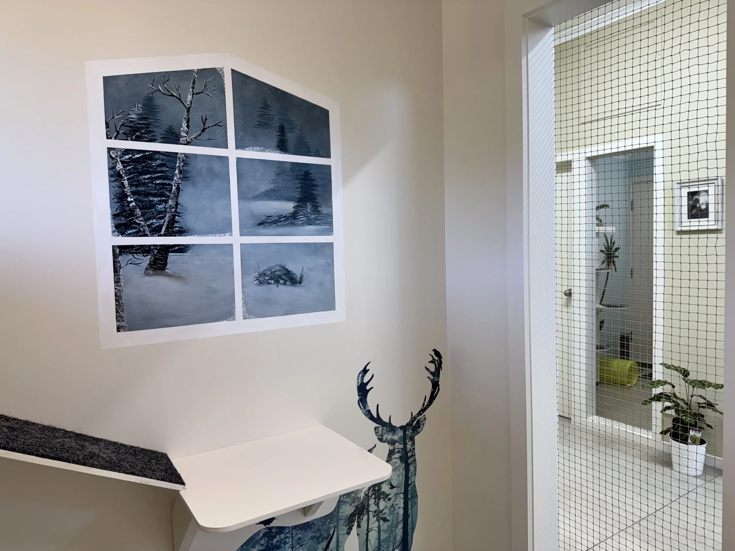 A cozy room with a decorative wall art of six black-and-white winter landscape photos, and a large blue deer silhouette decal. There is a small white shelf mounted on the wall and a office with potted plants visible through a door with a wire mesh grid.