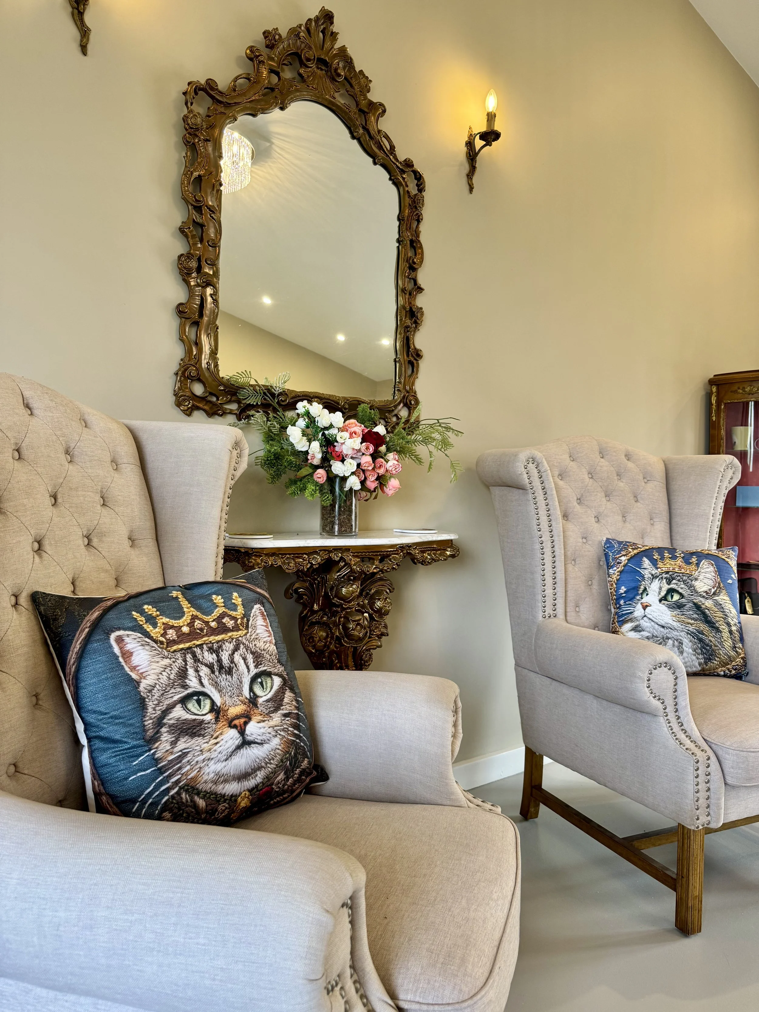 Elegant living room corner with a beige tufted armchair and a high-back beige armchair, each with decorative cat-themed pillows, a ornate mirror above a floral arrangement on a carved wooden console table, and a wall sconce lighting