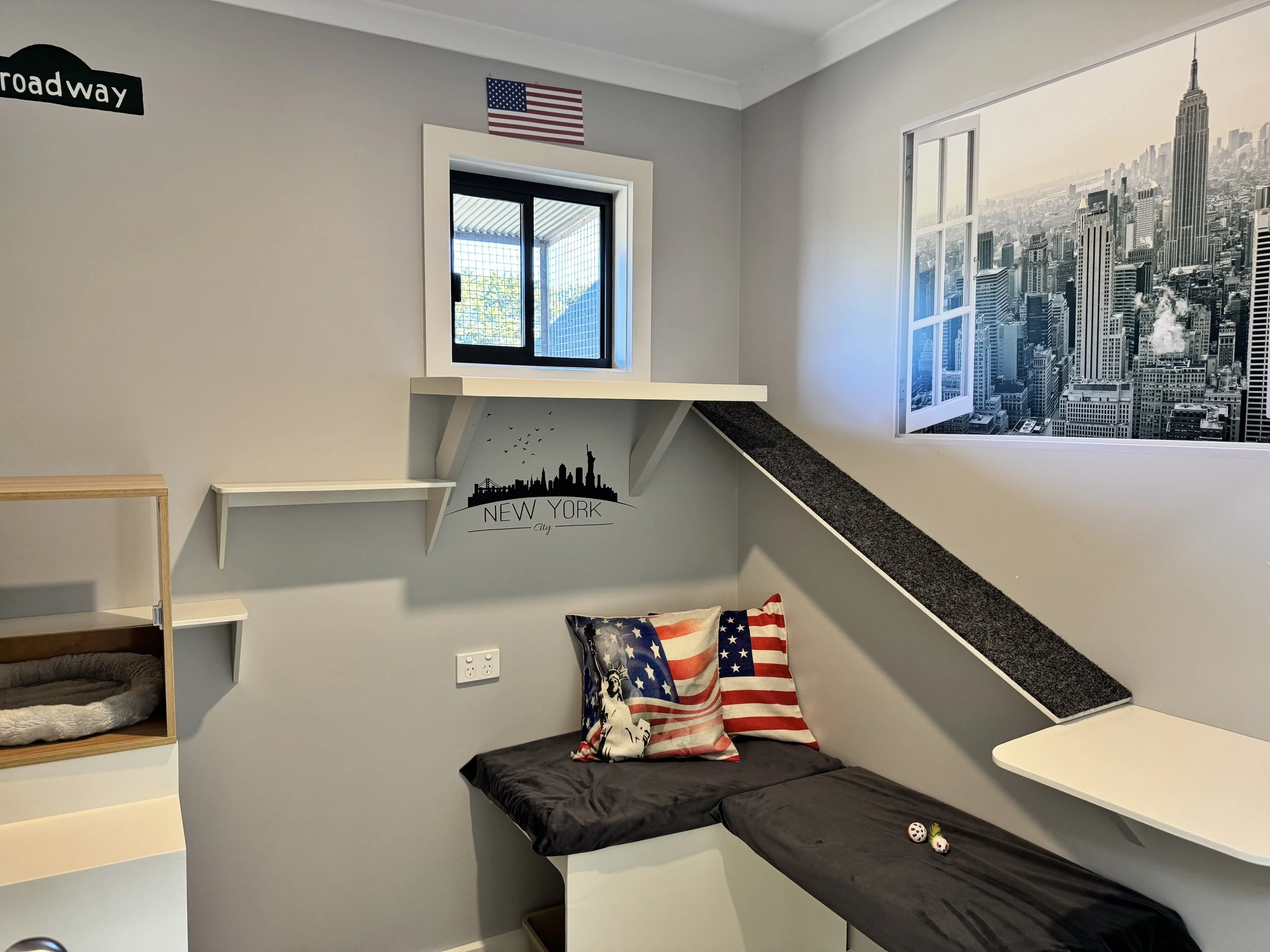 A corner of a room decorated with New York City and American flag-themed items, featuring a window look-out, a black and white cityscape wall decal, and American flag pillows on a black cushioned seat.