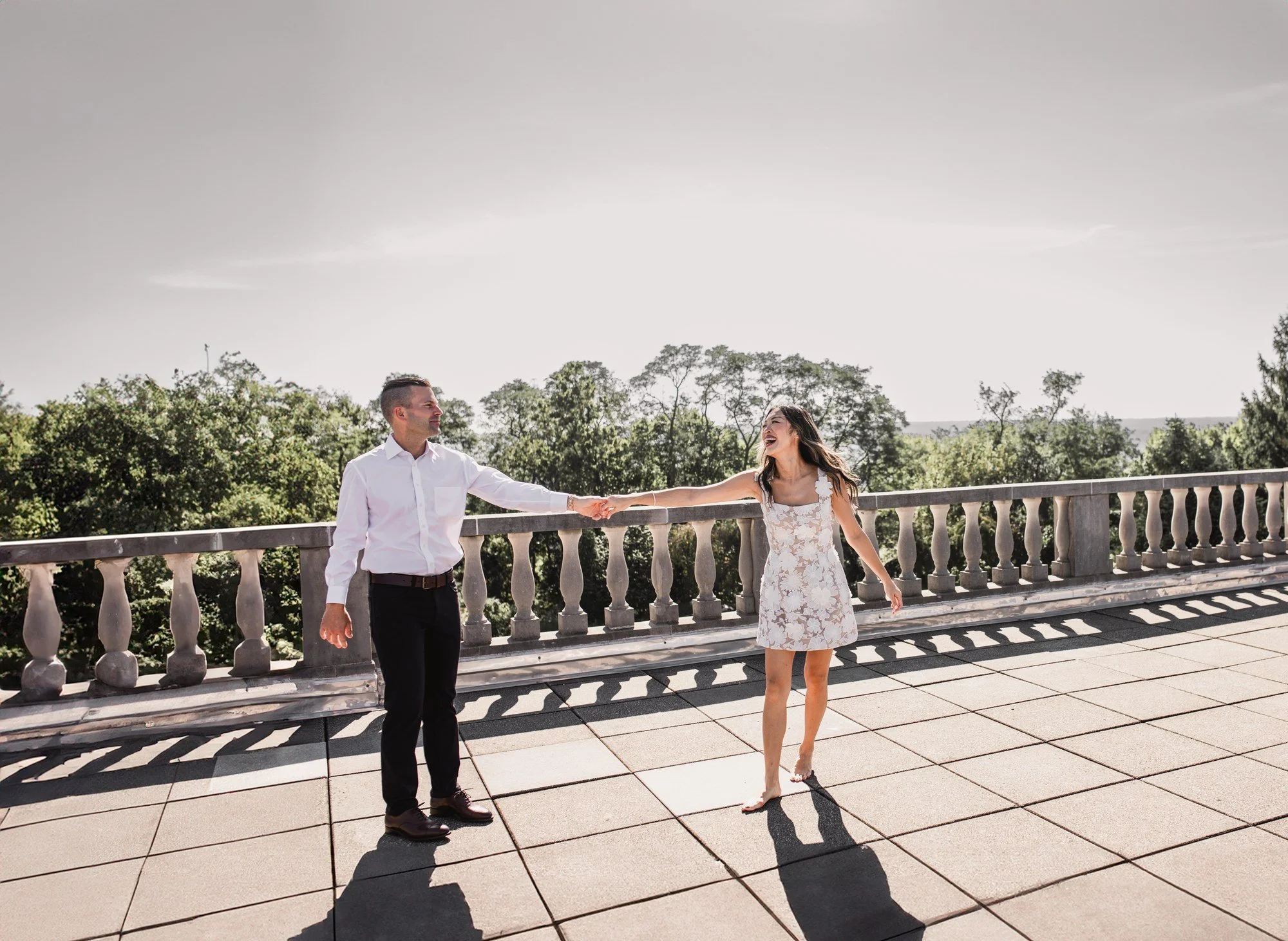 Kitty and Scott holding hands across a sunlit terrace with a stone balustrade and trees behind them.