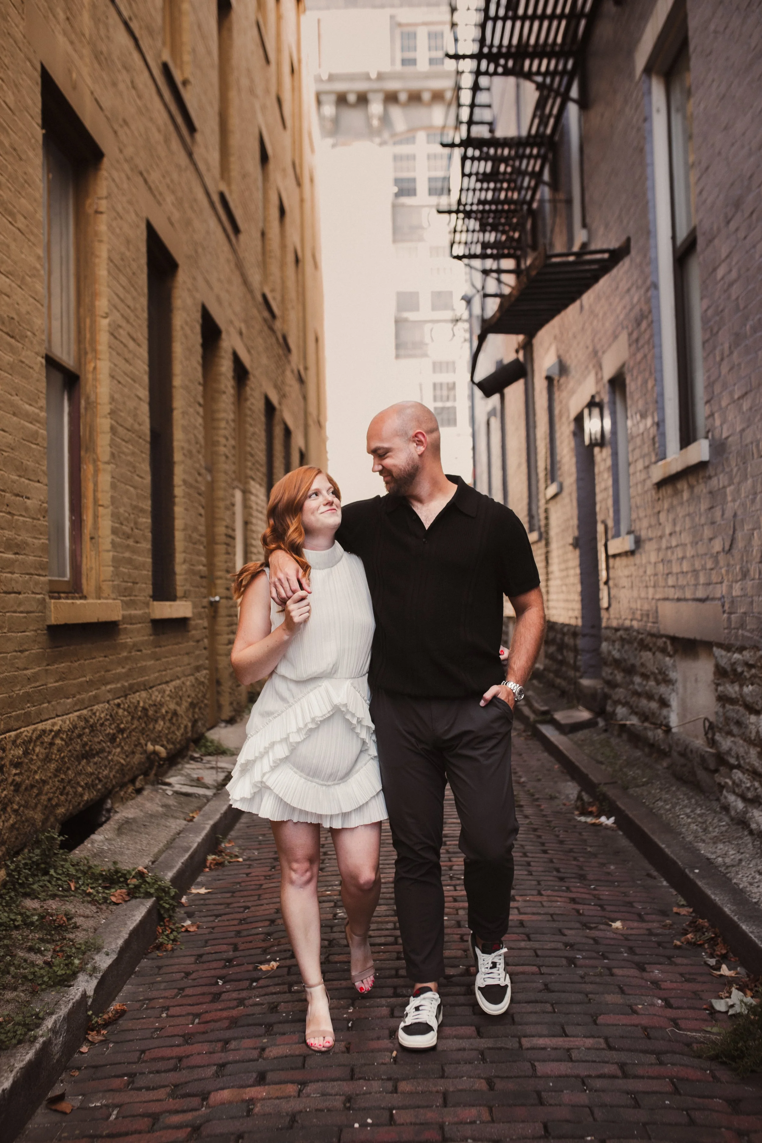 Engaged couple walking arm in arm through a brick alley and smiling at each other.
