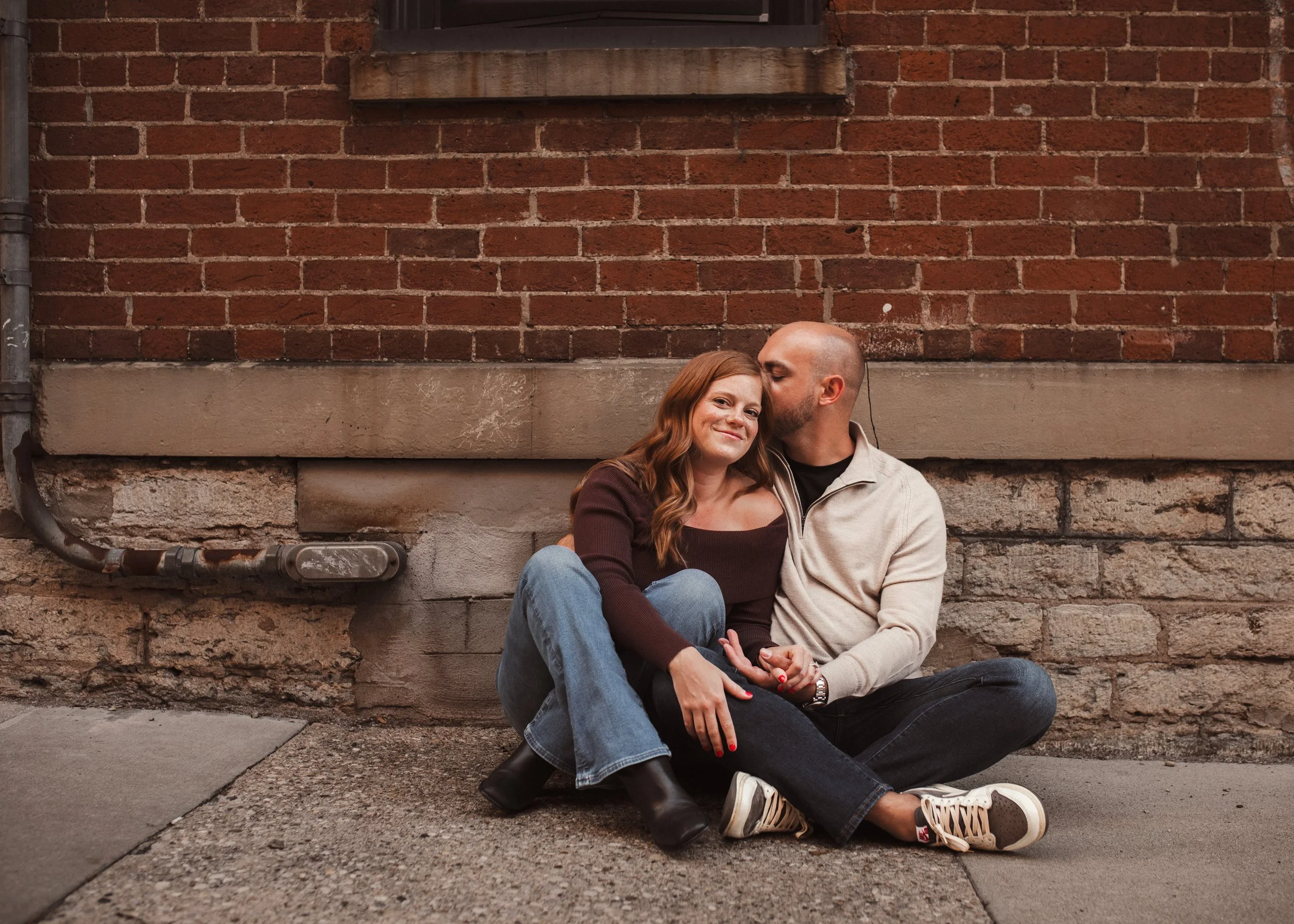 Engaged couple sitting close together beside a brick wall as one kisses the other on the head.