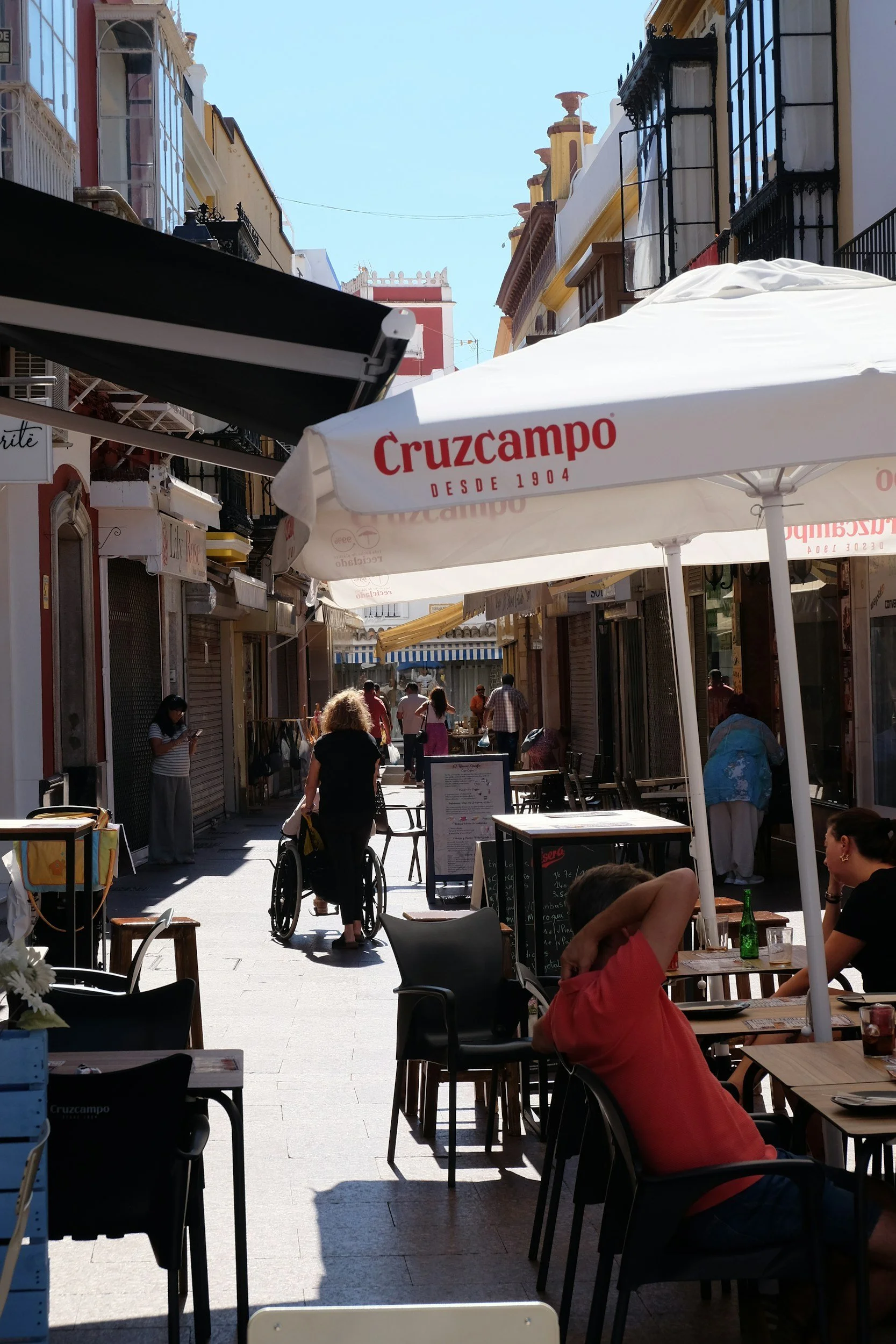 Outdoor café on a narrow pedestrian street with white Cruzcampo umbrellas, people at tables, and a wheelchair being pushed down the street.